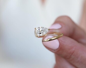 Könnte beinhalten: Ein goldener Ring mit einem birnenförmigen Diamanten. Das Band des Rings hat ein fließendes Design, und der Diamant funkelt. Der Ring wird vor einem weichen Hintergrund gehalten, um sein elegantes Design hervorzuheben.