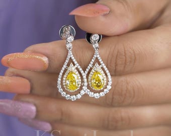 May include: A pair of elegant drop earrings featuring a pear-shaped yellow gemstone center, surrounded by a halo of smaller, clear gemstones. The earrings have a delicate, openwork design and are set in a silver-colored metal. The text "KSentJewel" is visible at the bottom.
