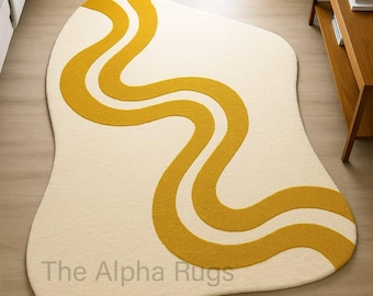 May include: Cream-colored, free-form rug with a mustard yellow, wavy design. The rug has a soft, textured appearance and is placed on a light wood floor. The text "The Alpha Rugs" is visible on the bottom of the rug.