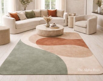 May include: A modern living room featuring a large, cream-colored sectional sofa with decorative pillows. A round, stone coffee table sits on a large area rug with a green, rust, and beige abstract design. The Alpha Rugs logo is visible.
