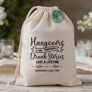 May include: A beige drawstring bag with the text "Hangovers are Temporary, Drunk Stories Last a Lifetime" in black script. The bag also includes the text "Sabrina's Last Trip" and a green circular sticker with text.