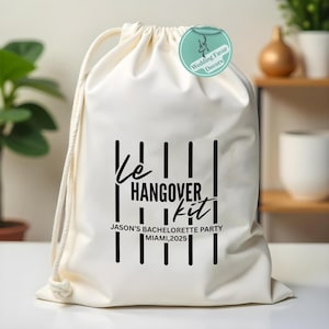 Pode incluir: Uma bolsa branca com cordão, com linhas verticais pretas e a frase "Le HANGOVER kit". Abaixo, lê-se "JASON'S BACHELORETTE PARTY MIAMI,2025". Uma etiqueta redonda com um veado está anexada.