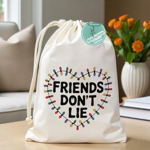 May include: Cream-colored drawstring bag with the text "FRIENDS DON'T LIE" inside a heart-shaped design of colorful string lights. A small, round tag with text is attached to the bag. The bag is sitting on a wooden surface.