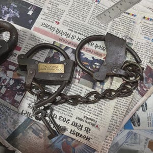 Puede incluir: Grilletes y candado de metal antiguo con el texto "ALCATRAZ SAN FRANCISCO" en una placa de latón. Los grilletes están conectados por una cadena. Varias llaves están al lado del candado. Sobre un fondo de periódico.