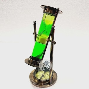 May include: An antique-style hourglass with a brass frame and a green-colored sand-like substance inside. The hourglass is mounted on a base that includes a compass. The design is reminiscent of nautical instruments.
