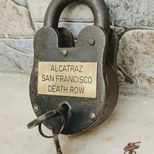 Könnte beinhalten: Ein verwittertes, dunkles Metallvorhängeschloss mit einem Schlüssel. Ein Messingschild auf dem Schloss trägt die Aufschrift "ALCATRAZ SAN FRANCISCO DEATH ROW". Das Schloss befindet sich vor einem strukturierten, hellfarbigen Hintergrund.