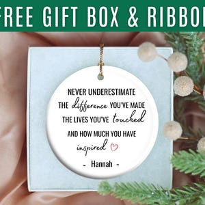 May include: A white ceramic ornament with gold string, inside a gift box. The ornament has the text "Never underestimate the difference you've made..." and is personalized with a name. A sprig of greenery and berries is in the background.