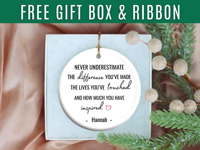 May include: A white ceramic ornament with gold string, inside a gift box. The ornament has the text "Never underestimate the difference you've made..." and is personalized with a name. A sprig of greenery and berries is in the background.