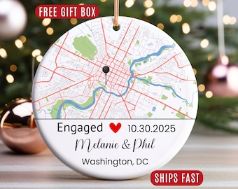 May include: A round white ceramic ornament featuring a map of Washington, DC with a black pin marker. Below the map, the text reads Engaged, a red heart symbol, 10.30.2025, the names Melanie & Phil, and the location Washington, DC.