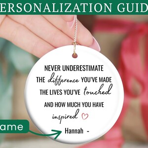 May include: A white ceramic ornament with a gold string, featuring the text "NEVER UNDERESTIMATE the difference you've made..." and a name. The ornament is held in front of a blurred background with gift boxes. The top banner reads "PERSONALIZATION GUIDE".