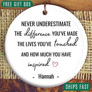 May include: A white ceramic ornament with a gold string for hanging. The ornament features the text "NEVER UNDERESTIMATE the difference YOU'VE MADE the LIVES YOU'VE touched AND HOW MUCH YOU HAVE inspired" and the name "Hannah".