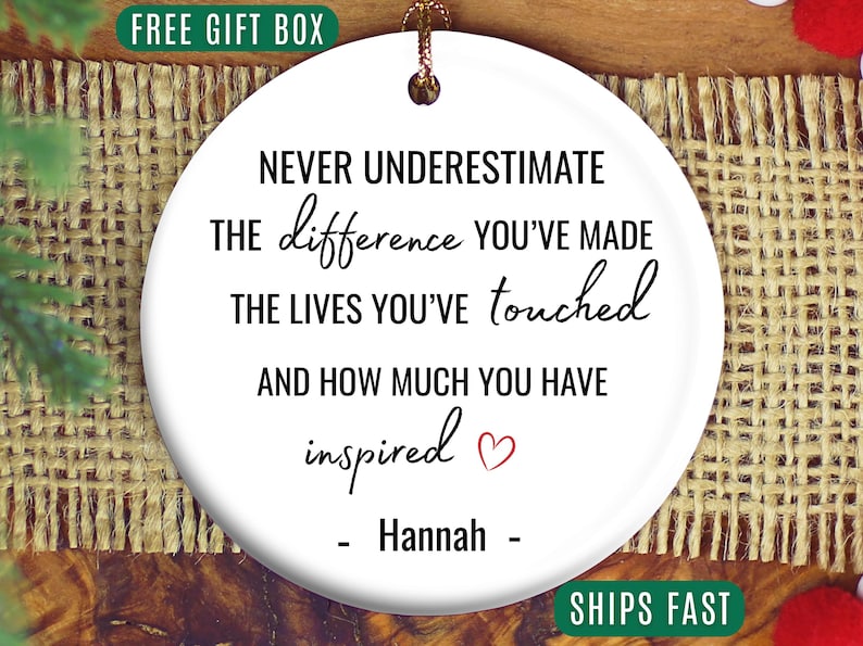 May include: A white ceramic ornament with a gold string for hanging. The ornament features the text "NEVER UNDERESTIMATE the difference YOU'VE MADE the LIVES YOU'VE touched AND HOW MUCH YOU HAVE inspired" and the name "Hannah".