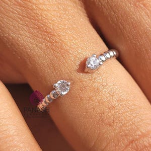 May include: A silver-toned open ring featuring a band of small, round, clear stones along each side, culminating in a single larger, round, clear stone at each of the two open ends. The metal band has a textured, beaded appearance.