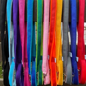 Puede incluir: Una colección de correas para perros de colores brillantes colgadas de una barandilla de madera. Las correas son de varios colores, incluyendo negro, azul, morado, rosa, verde, naranja, amarillo, gris y rojo. Cada correa tiene un mosquetón de metal.
