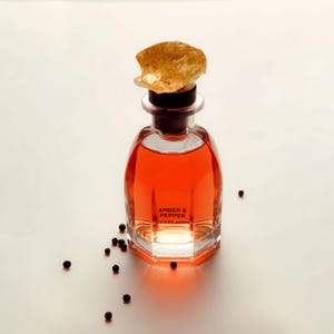 Puede incluir: Un frasco de perfume de vidrio transparente con base hexagonal lleno de líquido naranja. La etiqueta dice AMBER & PEPPER SISTER'S AROMA. El frasco está rematado con un tapón oscuro y una piedra grande, translúcida y de color ámbar. Hay granos de pimienta negra esparcidos cerca.