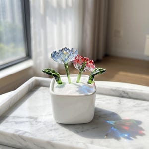 May include: A small white square ceramic planter filled with white sand, containing two glass flowers with green stems and leaves. One flower is light blue and the other is red, both featuring delicate, translucent petals and clear glass stems.