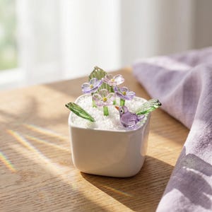 Könnte beinhalten: Ein kleiner, weißer quadratischer Keramiktopf gefüllt mit weißen Granulaten, Glasblumen und einem Glasvogel. Die Blumen sind lila und klar, mit grünen Stielen und Blättern. Der Vogel ist lila mit grünen Flügeln und einem gelben Schnabel.
