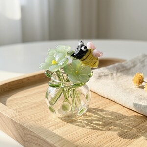 Puede incluir: Un pequeño jarrón de cristal transparente que contiene tres flores de cristal verde claro con centros amarillos y una figura de abeja de cristal con cuerpo a rayas amarillas y negras y pequeñas alas rosas posada sobre las flores como adorno.