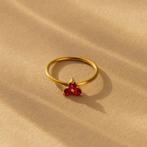 May include: A gold ring featuring a cluster of three small, round, vibrant red gemstones arranged in a triangular floral pattern. The band is thin and polished, showcasing a smooth metallic finish that complements the bright red stones.