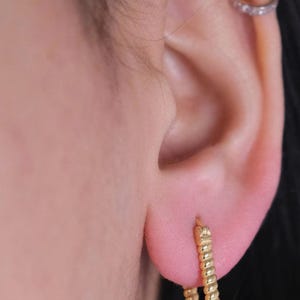 May include: Close-up of a gold hoop earring with a textured, twisted design. The earring is worn on a person's ear, with a small, delicate gold ring with small stones in the upper ear. The skin is light-toned.