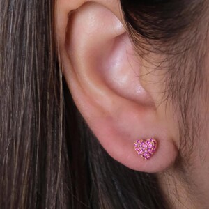 May include: A small heart-shaped stud earring featuring a gold-tone metal setting encrusted with numerous tiny, vibrant pink stones. The earring is positioned on the earlobe, showcasing a compact design with a textured, sparkling surface.