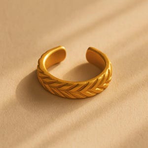 May include: A gold-colored adjustable ring with a textured, leaf-like pattern. The open-ended design allows for size adjustment. The ring is made of a shiny, metallic material.