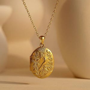 May include: A gold-toned oval locket pendant suspended from a matching delicate link chain. The locket features an embossed design on its front surface, depicting a bird perched among stylised floral and leafy botanical elements.