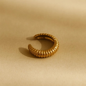 May include: A gold-colored ear cuff with a ribbed design. The open-ended cuff curves in a crescent shape, with a textured surface. The jewelry piece is set against a neutral background, highlighting its metallic sheen.