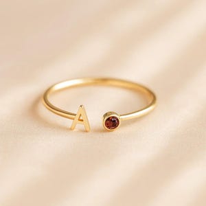 May include: A gold-toned open ring featuring a capital letter A on one end and a small, round, deep red gemstone set in a bezel on the other end. The band is thin and smooth, creating a delicate and minimalist appearance for this piece of jewelry.