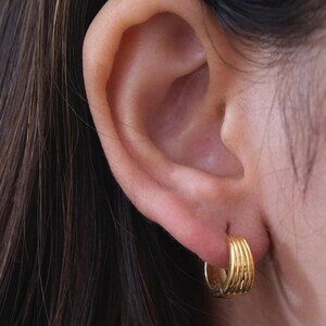 Puede incluir: Pendientes de aro dorados con un diseño texturizado y en capas. Los pendientes son pequeños, circulares y tienen un acabado brillante. Los pendientes se llevan en una oreja.