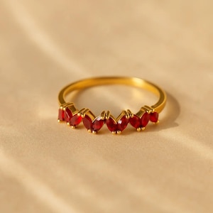 Puede incluir: Un anillo dorado con una hilera de piedras preciosas rojas en forma de marquesa. Las piedras están engastadas en la parte superior del anillo, creando un patrón continuo. El anillo se muestra sobre una superficie neutra y clara.