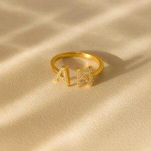 Pode incluir: Um anel dourado com as letras "A" e "N", cada uma incrustada com pequenas pedras preciosas transparentes. O anel é apresentado sobre um fundo macio e creme, realçando o seu design e brilho.