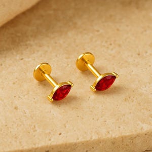Op de afbeelding: Twee goudkleurige labret-studs met een marquise-geslepen rode edelsteen in een klauwzetting. Elk sieraad bestaat uit een platte ronde achterkant, een rechte stift en een decoratief voorstuk met een felrode steen.
