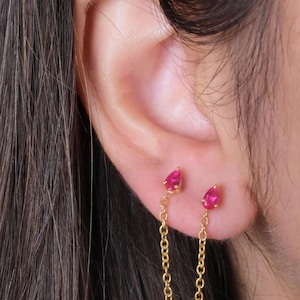 Pode incluir: Brincos pendurados dourados com uma pequena pedra preciosa rosa brilhante em forma de gota na parte superior e inferior. As pedras preciosas são conectadas por uma delicada corrente dourada. Os brincos são mostrados em uma orelha.