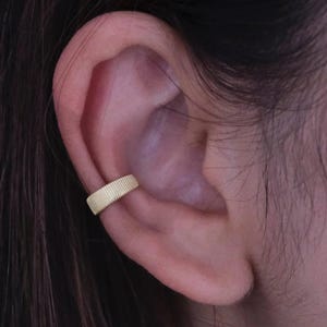 May include: A gold-colored ear cuff with a ridged texture is worn on the ear. The cuff is a simple band that wraps around the ear cartilage. The image shows the ear and the cuff in close-up.