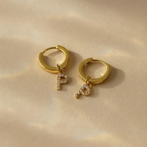 May include: A pair of gold-colored hoop earrings. Each earring has a small, dangling charm in the shape of the letter "P", encrusted with small, clear crystals. The earrings are displayed on a light beige surface.