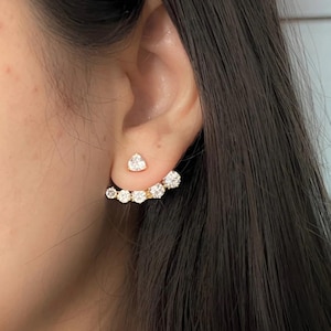 Puede incluir: Pendientes dorados con piedras claras y brillantes. Un pendiente tiene un perno en forma de corazón, el otro un diseño curvo con múltiples piedras. Los pendientes están sobre cabello oscuro.