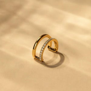 May include: A gold-colored ear cuff with a double-band design. One band is solid, while the other is adorned with small, sparkling gemstones. The jewelry piece is set against a neutral background, highlighting its elegant form.