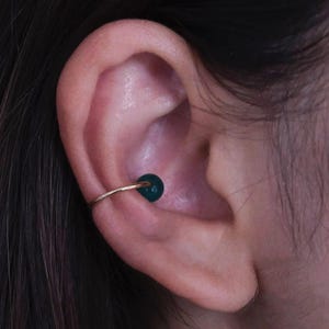 May include: A gold-colored ear cuff with a dark green, round bead. The cuff is worn on the ear's cartilage, with the bead positioned on the inner ear. The jewelry is simple and elegant.