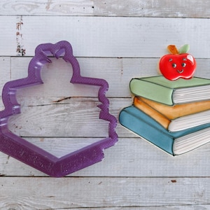 Peut inclure: Emporte-pièce en plastique violet en forme de pile de livres. À côté se trouve un biscuit décoré d'une pile de livres surmontée d'une pomme rouge. La pomme a un visage souriant.