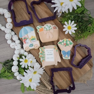 May include: A wooden cutting board displays decorated sugar cookies and purple cookie cutters. The cookies are shaped like a flower cart, bouquet, veggie bag, and a frame with the text "She's Fresh Off The Market July 11, 2015."