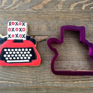 May include: A red and black cookie cutter shaped like a vintage typewriter with a paper insert that says "XOXO" in red letters. The cookie cutter is next to a red and black cookie decorated with white dots and the same "XOXO" paper insert.