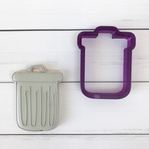 May include: A grey cookie in the shape of a bin with a handle. Next to it is a purple plastic cookie cutter in the shape of a bin.