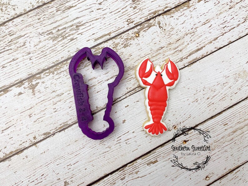 May include: Purple plastic lobster cookie cutter with the text "Crafter 2nd" on the side, next to a red and white iced lobster cookie.