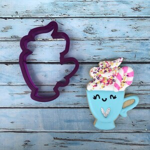 Puede incluir: Una galleta azul y blanca con forma de taza con crema batida, chispas y un bast&oacute;n de caramelo en la parte superior. La galleta tiene una carita linda con ojos y un coraz&oacute;n. Tambi&eacute;n se ve un cortador de galletas de pl&aacute;stico morado con forma de taza en la imagen.