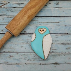 May include: A blue and white owl-shaped cookie on a blue wooden background. A wooden rolling pin is in the background.