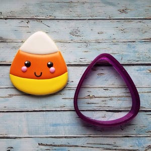 May include: A purple candy corn cookie cutter with a yellow, orange, and white candy corn cookie next to it.