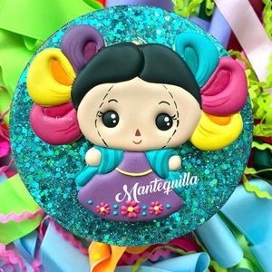 May include: A round, teal glitter-covered ornament with a colorful, cartoon-style doll wearing a purple dress and a turquoise jacket. The doll has black hair and large eyes. The ornament has the text "Mantequilla" written on it.