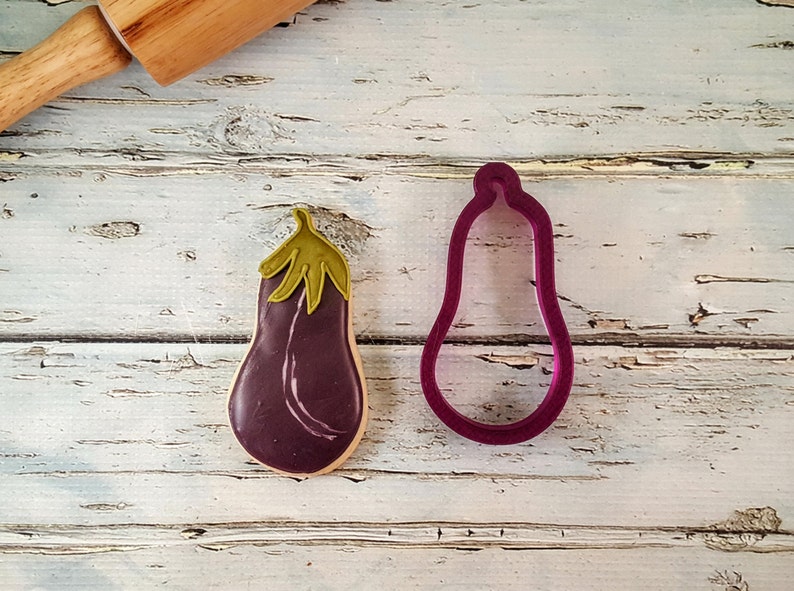 May include: A purple eggplant cookie cutter and a decorated eggplant cookie on a white wooden background.