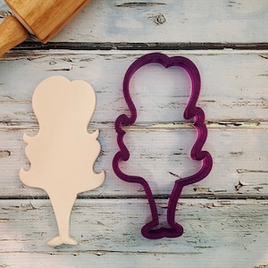 May include: Two cookie cutters, one is a light brown color and the other is a dark purple color. Both cutters are shaped like a person with long hair.